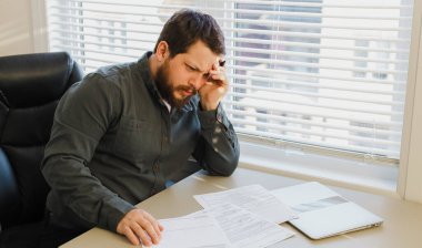 Ofisteki belgelerle çalışan üzgün bir yönetici, masadaki bilgisayarla kapalı..