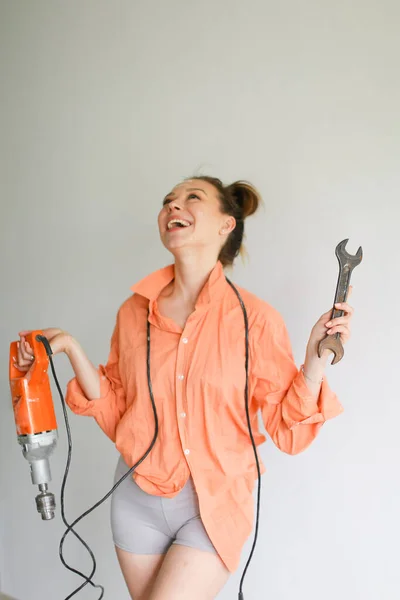 Joven chica sonriente sosteniendo un taladro y una llave inglesa, usando una camisa naranja de pie sobre un fondo aislado. —  Fotos de Stock