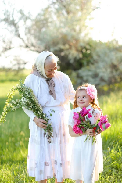 Velha avó de pé com pouco bonito neta e mantendo flores. — Fotografia de Stock