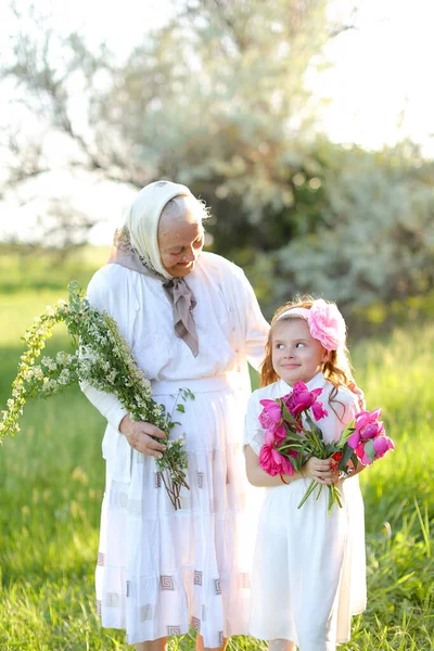 Velha avó de pé com pouco bonito neta e mantendo flores. — Fotografia de Stock