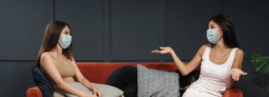 İki kadın evde oturuyor ve koruyucu maske takıyor.