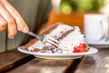 Açık kahve masasında bir dilim çikolatalı ve çilekli kek yiyen birinin yakın çekimi.