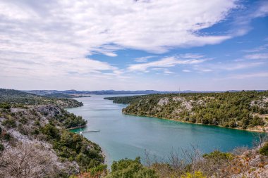 Hırvatistan 'daki Krka nehrinin turkuaz su ve mavi gökyüzünün altındaki yeşil tepelerle manzarası