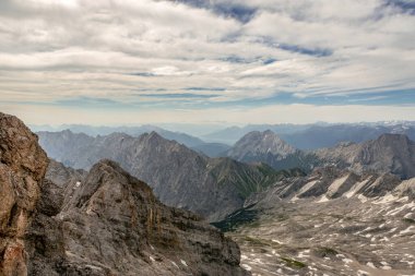 Yazın Zugspitze yakınlarındaki dağların etkileyici manzarası. Engebeli kayalar, kar bantları ve uzak vadiler parçalı bulutlu bir gökyüzünün altında çarpıcı bir dağ manzarası oluşturur..