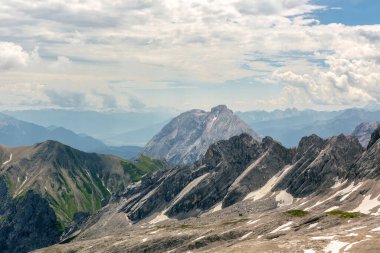 Yazın Zugspitze yakınlarındaki dağların etkileyici manzarası. Engebeli kayalar, kar bantları ve uzak vadiler parçalı bulutlu bir gökyüzünün altında çarpıcı bir dağ manzarası oluşturur..