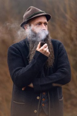 Uzun sakallı ve geleneksel şapkalı olgun bir adam sessiz bir ortamda pipo içiyor. Doğal ışık ve sisli arka plan sakin, yansıtıcı bir sonbahar havası yaratır. Sahnenin kırsal ve düşünceli karakterini vurgular..