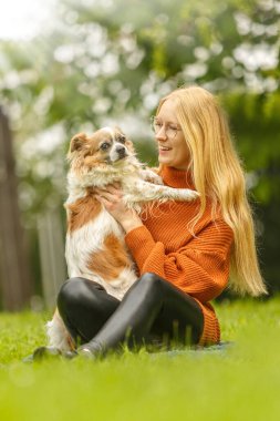 Uzun sarı saçlı ve gözlüklü gülümseyen genç bir kadın uzun saçlı Chihuahua 'sını kollarında tutuyor. Yeşil yapraklar ve beyaz çiçeklerle yemyeşil bir bahar bahçesinde dururlar, sıcaklık ve şefkat yayarlar..