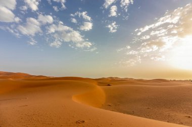 Fas 'taki Erg Chebbi çölünün kum tepelerinin üzerinde, bulutlu bir gökyüzünün altında sıcak akşam ışığı.
