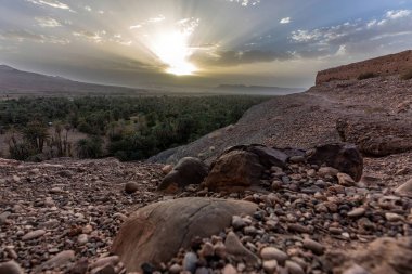 Fas vahasını aydınlatan altın gün batımı ve ön planda dağılmış kayalarla çöl tepeleri.