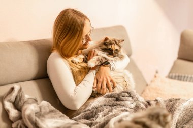 Sıcak bir iç mekan sahnesinde, yumuşak battaniyelerle kanepede uzun tüylü kedisini kucaklayan gülümseyen bir kadın.