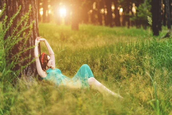 Красивая молодая женщина в элегантном светло-зеленом платье лежит под деревом в лесу с лучами солнечного света. Тонирование — стоковое фото