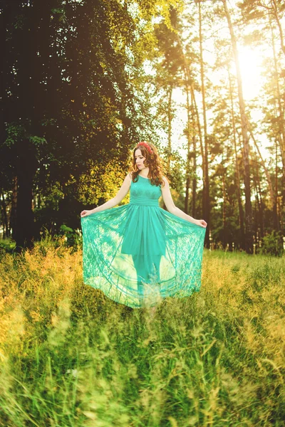 Mulher bonita vestindo elegante vestido verde claro em pé na floresta com raios de luz solar irradiando através das folhas das árvores. Foco suave e Tonificação — Fotografia de Stock