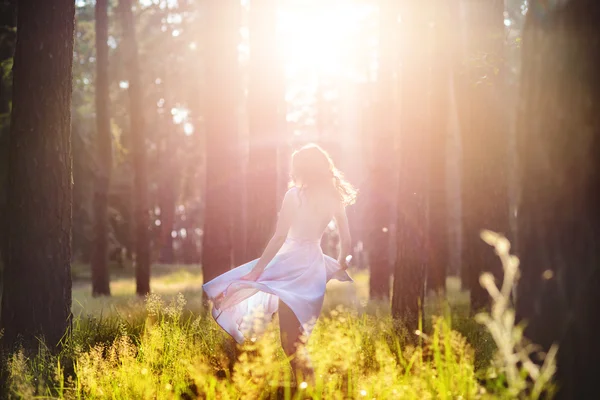 Belle jeune femme portant une élégante robe bleu clair debout dans la forêt avec des rayons de soleil rayonnant à travers les feuilles des arbres. Mise au point douce et tonification — Photo