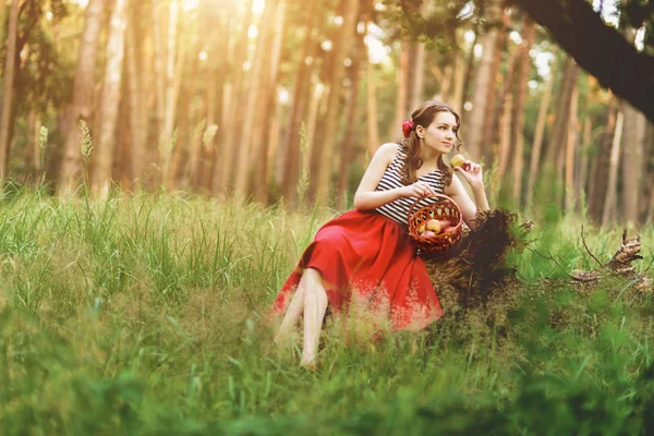 Menina sentada na floresta com uma cesta de maçãs em uma saia longa vermelha — Fotografia de Stock