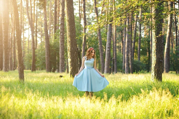 Bella giovane donna che indossa elegante abito azzurro in piedi nella foresta con raggi di luce solare che irradia tra le foglie degli alberi. Tonificante — Foto Stock