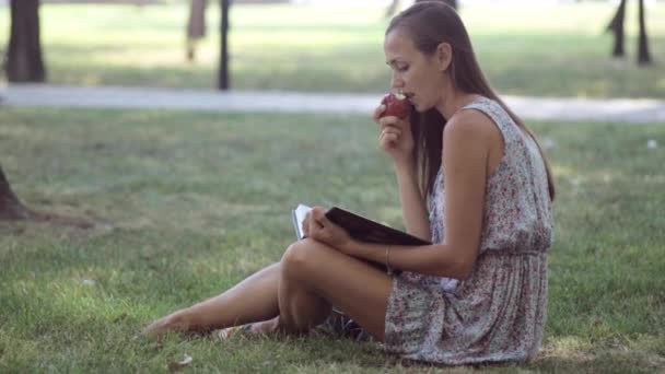 Fiatal nő olvasó könyv és étkezési Alma városban park. — Stock videók