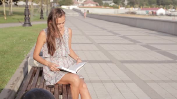 Молодая женщина сидит на скамейке с книгой — стоковое видео