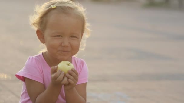 Ένα χαριτωμένο μικρό κορίτσι τρώει ένα φρέσκο μήλο στο πάρκο και να δείχνει το μεγάλο δάχτυλο μέχρι. — Αρχείο Βίντεο