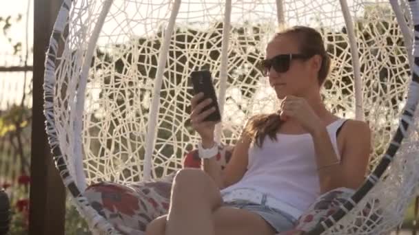 Young woman sitting on the swing and click cellphone at sunset — Stock Video