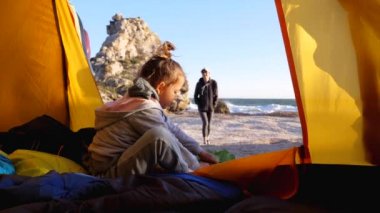 Kumsalda kamp çadırında otururken annesi kızının ayakkabılarını giymesine yardım ediyor.