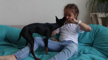 Küçük kız kanepede siyah oyuncak teriyer köpeğiyle oynuyor. 