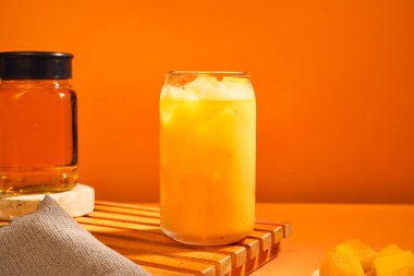 Bir bardak mango suyu, buz küpleri ve turuncu arka planda bir şişe su..