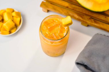 Mango suyunun üst açısı şeffaf bardağın içinde buz küpleri ve bir parça mango meyvesi ve gri peçeteyle..