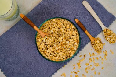 Yuvarlak altın yulaflarla dolu cam bir kâse, ahşap kaşıkların yanına desenli mor bir kumaş ve kahvaltıya hazır bir bardak süt..