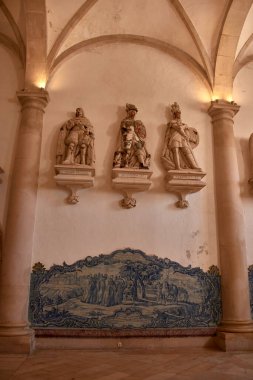 The chapter house of the Alcobaca Monastery preserves Its solemn atmosphere features Baroque sculptures depicting crowned religious figures, arranged on a stone wall with decorative inscriptions.