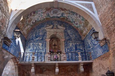 The Porta da Vila in Obidos is presented as a monumental entrance with a stone arch framing a niche decorated with blue tiles, floral frescoes, and a small altar with a cross and flowers. 