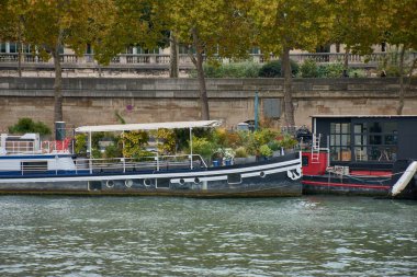 Paris 'teki Seine Nehri boyunca bir yüzen ev mavnası, yeşillik ve kentsel tasarımı birleştiren bir bahçe çatısı ile demirlidir. Sahnede sonbahar ağaçları, bir taş duvar ve nehir kenarında bir gezinti alanı yer alıyor. Huzurlu ve pitoresk bir sahne yaratıyorlar.. 