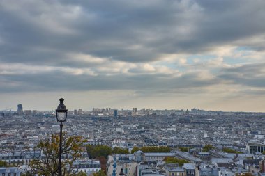Montmartre tepesinden, Paris 'in panoramik manzarası, gri çatılar, dar sokaklar ve ufka kadar uzanan kentsel bir kumaşı gözler önüne seriyor. Parçalı bulutlu gökyüzü derinlik ve dinginlik ekler, etrafındakiler ise bohemleri çağırır. 
