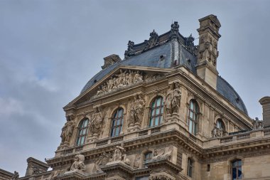 Louvre Müzesi 'nin ön cephesi, geçmişini kraliyet sarayı olarak andıran sütunlar, heykeller ve kemerlerle işaretlenmiş klasik mimarisine dikkat çekiyor. Bulutlu gökyüzü sahneye dram eklerken, simetri dizginleri...