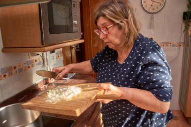 71 yaşında bir kadın pijamalarıyla vejetaryen yemeği hazırlıyor, ev mutfağında tahta bir tahtada sebze doğruyor. Ilık ışıklandırma, canlı renkler ve günlük ortam sahnenin gerçekliğini pekiştiriyor.