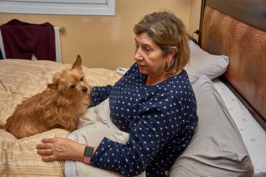 71 yaşındaki bir kadın, egzersize gitmeden önce makyajını odaklayarak, dudaklarına renk katarak ve gözlerini kapayarak yapar. Hala pijama giyiyor, sıcak ve basit bir ev ortamında enerji, kendine güven ve aktif yaşlanma yayıyor..