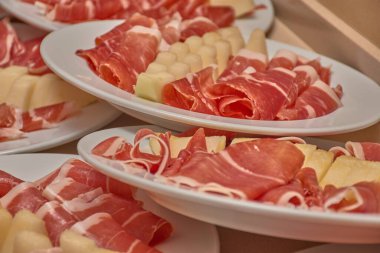 Düzinelerce beyaz tabakta, büyük bir toplantı için sıra sıra dizilmiş kavun dilimleri eşliğinde pişirilmiş jambon kanepeleri bulunur. Düzenli kompozisyon ve jambonun pembesi ile kavunun soluk sarısı arasındaki ahenkli renk düzeni