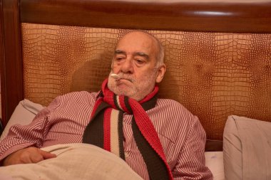 Yaşlı bir adam, ateşi ağzında bir termometreyle ölçülürken yatakta yatar. Geleneksel kumaşlarla süslenmiş sıcak bir odada örülmüş bir battaniyeyle kaplı çizgili pijama ve kırmızı bir eşarp giyiyor.. 