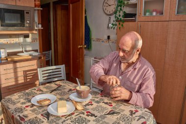 Yaşlı bir adam, güneş ışığı pencereden içeri akarken ve masayı yıkarken mutfakta huzurlu bir kahvaltı yapıyor. Çizgili bir gömlek giyiyor ve etrafı kahve ile çevrili bir tostu yağlamaya odaklanıyor, tatlım.