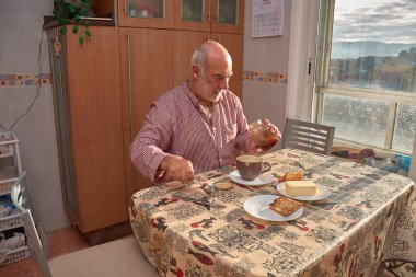 Yaşlı bir adam, güneş ışığı pencereden içeri akarken ve masayı yıkarken mutfakta huzurlu bir kahvaltı yapıyor. Çizgili bir gömlek giyiyor ve etrafı kahve ile çevrili bir tostu yağlamaya odaklanıyor, tatlım.