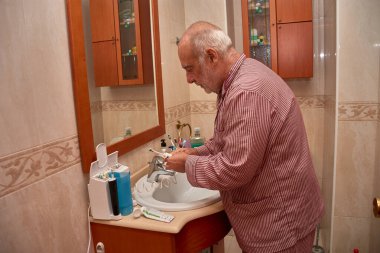 Çizgili pijama giymiş yaşlı bir adam elektrikli diş fırçasıyla dişlerini fırçalarken lavabonun üzerine eğildi. Banyoda dekoratif kenarlı bej fayanslar, ahşap mobilyalar ve tezgahın üzerinde çeşitli hijyen ürünleri bulunuyor.. 