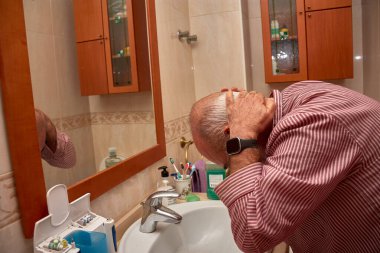 Kırmızı ve beyaz çizgili gömlek giyen yaşlı bir adam lavaboda tımar rutinini yaparken küçük bir şişe tutuyor. Banyoda dekoratif kenarı olan bej fayanslar, büyük bir ayna ve cam kapılı ahşap dolaplar bulunuyor.. 