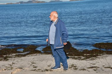 Yaşlı bir adam güneşli bir günde kumsalda yürür, mavi ceket, siyah pantolon ve düz bir şapka giyer. Sahil manzarasının yumuşak ışığı ve sakin deniz huzur ve refahın atmosferini yükseltir.. 