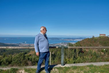 Yaşlı bir adam güneşli bir günde Monte Alba 'dan Vigo Estuary' yi düşünürken tahta korkuluklara yaslanır. Mavi gökyüzü, berrak ışık ve arka planda binalar ve dağlar olan kıyı manzarası dinginlik atmosferini güçlendirir. 