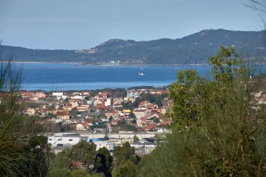 San Andres de Comesana tepelerinden görülen Ria de Vigo 'nun yüksek manzarası, konut binaları, canlı bir haliç ve açık bir gökyüzü içerir. Görüntü, kentsel gelişim ve doğa arasındaki uyumu yakalıyor ve sahil şeridini gösteriyor.