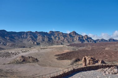 Teide Dağı 'nın görkemli zirvesinin ihtiyatlı bakışları altında Llano de Ucanca' nın tortulu ovasından yoğun kara lav akıntıları geçer. Doğrusal bileşim, yerin engebeli dokusundan mavi ufka kadar gözü yönlendirir.