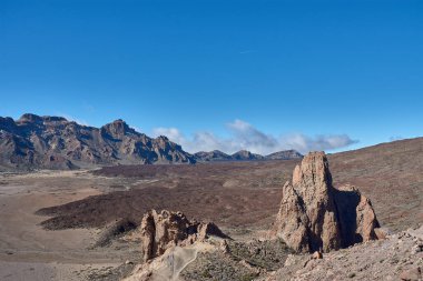Teide Dağı 'nın görkemli zirvesinin ihtiyatlı bakışları altında Llano de Ucanca' nın tortulu ovasından yoğun kara lav akıntıları geçer. Doğrusal bileşim, yerin engebeli dokusundan mavi ufka kadar gözü yönlendirir.