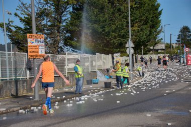 Nigran, Pontevedra, İspanya; 6 Nisan 2025: Yüksek görüşlü yelek giyen genç gönüllüler İspanya 'nın Nigran kentinde düzenlenen Vig Bay Half Marathon sırasında koşuculara suya batırılmış sünger dağıtıyorlar. Sahne, toplum desteği ve ekip çalışmasının bir anını partik olarak yakalıyor.