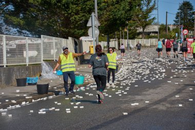 Nigran, Pontevedra, İspanya; 6 Nisan 2025: Yüksek görüşlü yelek giyen genç gönüllüler İspanya 'nın Nigran kentinde düzenlenen Vig Bay Half Marathon sırasında koşuculara suya batırılmış sünger dağıtıyorlar. Sahne, toplum desteği ve ekip çalışmasının bir anını partik olarak yakalıyor.