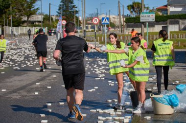 Nigran, Pontevedra, İspanya; 6 Nisan 2025: Yüksek görüşlü yelek giyen genç gönüllüler İspanya 'nın Nigran kentinde düzenlenen Vig Bay Half Marathon sırasında koşuculara suya batırılmış sünger dağıtıyorlar. Sahne, toplum desteği ve ekip çalışmasının bir anını partik olarak yakalıyor.