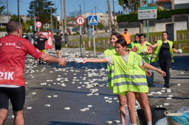 Nigran, Pontevedra, İspanya; 6 Nisan 2025: Yüksek görüşlü yelek giyen genç gönüllüler İspanya 'nın Nigran kentinde düzenlenen Vig Bay Half Marathon sırasında koşuculara suya batırılmış sünger dağıtıyorlar. Sahne, toplum desteği ve ekip çalışmasının bir anını partik olarak yakalıyor.